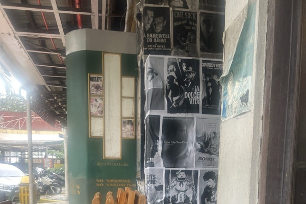 The café pillar at EXT.Cafe, papered in black-and-white film posters, with a yellow armchair in front.